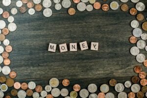 A wooden surface bordered by scattered coins, with Scrabble tiles spelling the word "MONEY" in the center