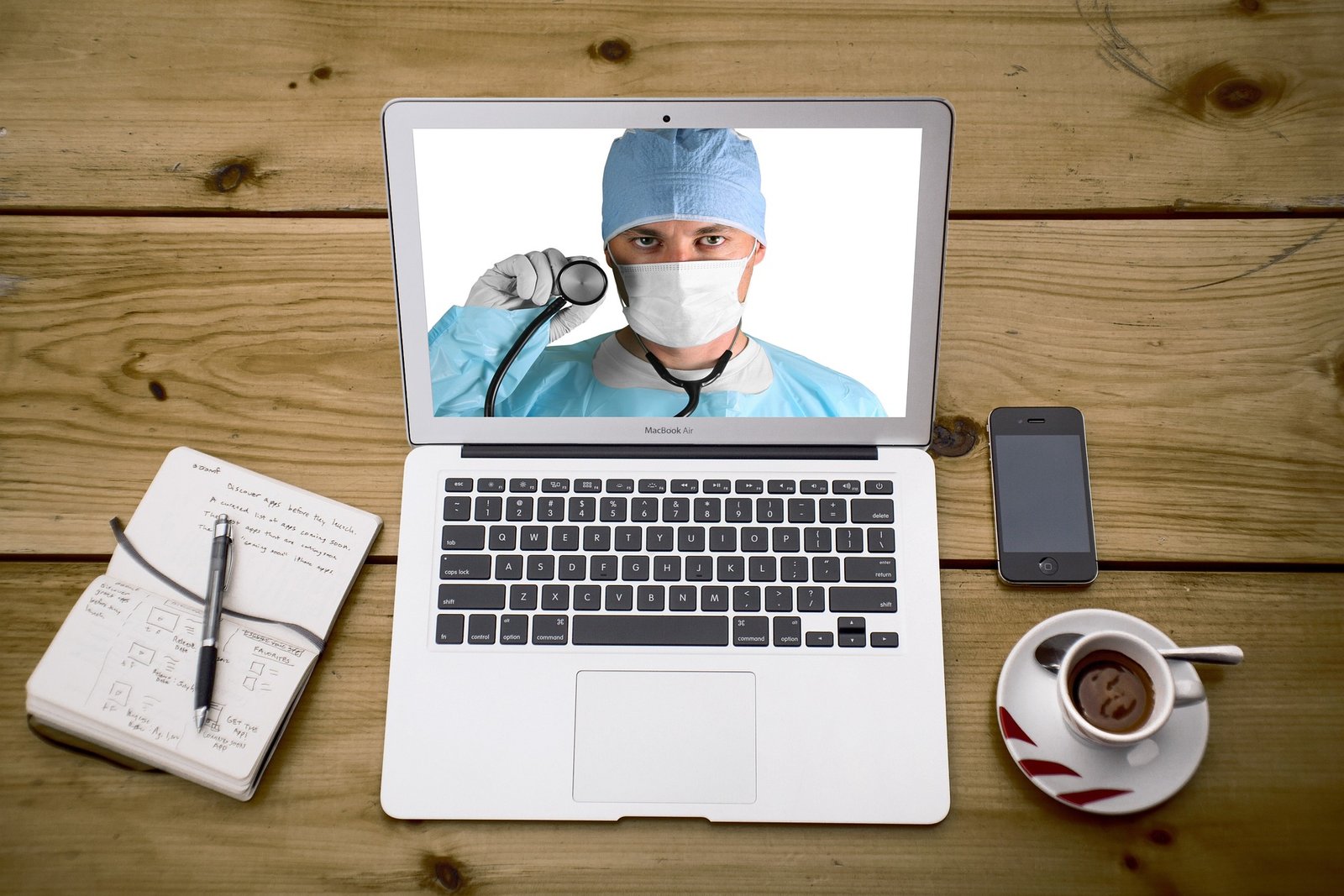 A laptop screen displaying a doctor with a stethoscope, symbolizing telemedicine, surrounded by a notebook, smartphone, and coffee cup.