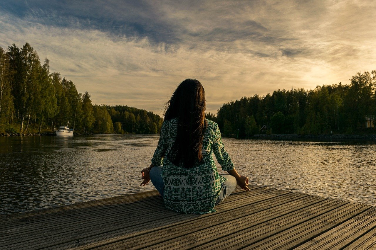 A woman meditating on a wooden dock by a peaceful lake during sunset.